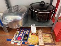 Photo showing vintage aluminum Wagner Ware Sidney O Magnalite oval Dutch oven roaster with lid, black Crock-Pot slow cooker, and a collection of vintage cookbooks stacked beneath them on a shelf.