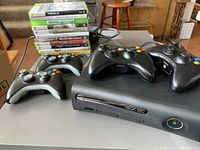 Xbox 360 console front and four controllers on table with game cases in background.