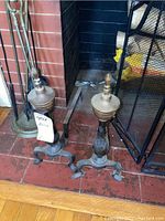 Pair of metal fireplace andirons set on tiled hearth next to fireplace screen, both showing signs of use and dirt accumulation.