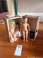 Three antique 10-inch Vogue dolls arranged in and near the carrying case on a wooden floor, showing condition and missing right arm on one doll.