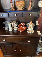 Dark wooden dresser with nine decorative items including two orange football-shaped bowls, clear crystal vase, white bowels, ceramic teapot with floral pattern, small white vase, and ceramic figurine of woman