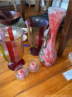 Photo showing two large multicolor striped glass vases, tall red and white speckled face vase, two small ribbed glass candle holders with pink candles on wooden floor.