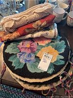 Six carpet floral chair pads stacked with lace and floral fabric table cloths under them on a table, surrounded by mugs and other items.