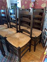 Six wooden dining chairs with ladder back design and reed woven seats arranged together in a room with floral wallpaper and wooden floor. Visible signs of wear including scratches on wooden frames.