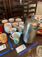 Photo showing one metal coffee pot and nine assorted ceramic mugs with different patterns and colors grouped on a wooden table.