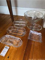 Photo showing six clear cut and pressed glass platters and dishes arranged on floor under table, varying sizes and shapes