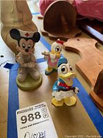 Three Disney figurines displayed on a blue and beige surface with wooden items in the background; Minnie Mouse dressed as a nurse, Donald Duck, and Daisy Duck playing musical instruments