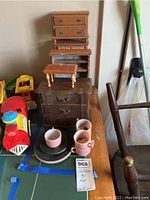 View showing two smaller wooden toy dressers stacked on top of a dark wood miniature armoire with a few small plates and cups on the armoire. A toy train engine is visible next to the furniture.