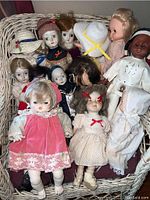 Wide shot of the lot showing all 13 dolls in a basket, mixed porcelain and plastic dolls with various clothing and hats