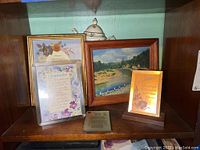 Shelf showing multiple framed items including inspirational pictures about grandmother, signed landscape painting, ceramic teapot on top shelf