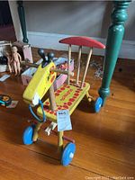 Front angle of vintage wooden Playskool giraffe scooter showing yellow and red paint, four blue wheels, and giraffe-shaped front with smiling face.