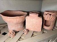 Five unglazed pottery pieces on wooden shelf; includes planter, vase, lantern, two pottery roses.
