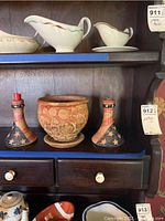 Shelf with two U.S. flag patterned pottery candle holders with red candle stubs and one round decorative pottery bowl or planter pot with textured circular patterns in earthy colors.