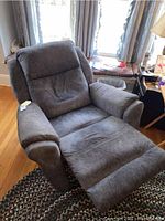Gray fabric reclining armchair in used condition with footrest extended, placed on a rug in front of windows with curtains.
