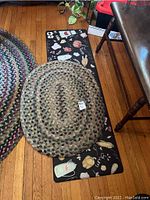 Two rugs shown on hardwood floor: oval braided rug on top of a rectangular cushioned rug with vegetable pattern.