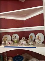 Wide view of eleven tea cups and five of twelve saucers displayed on shelves with a small brass bell.
