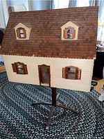 Front view of the vintage wooden dollhouse showing brown shingled hinged roof with two dormer windows and cream-colored walls with shuttered windows. Mounted on swivel wooden base.