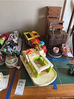 Photo of vintage toy boat with green and white deck, blue hull, showing wear and dirt. Includes other toys and small furniture in background.