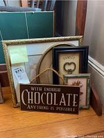 Six pieces of wall art stacked on floor showing variety of frame styles and sizes, including golden ornate frame and wooden plaque with chocolate quote.
