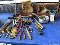 Full view of assorted hand tools laid out on a blue table including various hammers, pliers, screwdrivers, and tool holders.