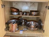 Cabinet shelf holding various metal mixing bowls, pots, and kitchen tools showing condition and variety of items.