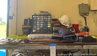 Photo of a workbench with various tools including drill bits, a white hard hat, a blue respirator mask, and other hand tools.