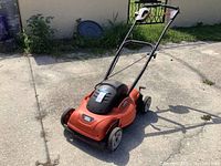 View of the lawn mower from the side showing handlebars, wheels, and mower body on concrete surface.