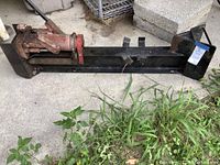 Full side view of the black metal frame and red hydraulic jack of the log splitter on concrete floor with grass nearby.