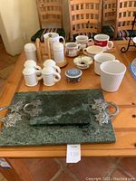 Photo shows two green marble cheese boards with pewter handles on table along with various white ceramic mugs, salt and pepper shakers, and other small containers grouped together.