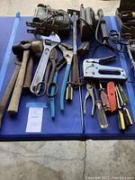 Overview of assorted hand tools laid out on table including hammers, wrenches, scissors, pliers, staple gun, screwdriver, utility knife, and hacksaw