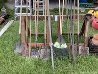 Full view of several flat shovels, spades, rake, and tree stakes with wooden handles and metal heads, standing in the grass and leaning against wooden barricades.