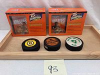 Front view of two Wheaties 75 Years of Champions collectible boxes and three hockey pucks displayed on a wooden tray. Each Wheaties box shows an athlete replica card with gold signature. Hockey pucks bear distinct team/logo designs.