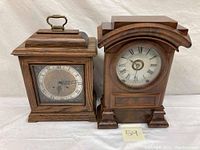 Two wooden mantle clocks side by side; one with brass handle and glass front, the other with arched top and round dial