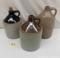 Front view of three stoneware jugs: beige tall jug, medium brown-glazed top jug, medium black-glazed top jug with cork stopper.
