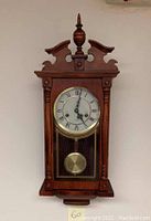 Full front view of the vintage wooden pendulum wall clock with decorative top and pendulum visible through glass panel.