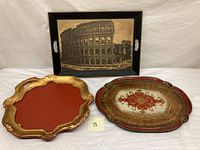 Three vintage trays displayed together, showing a black tray with a Colosseum print, red Florentine tole tray, and a large ornate Florentine tole tray