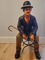 Front view of the Charlie Chaplin figurine seated on the cast iron bench, showing detailed facial features, clothing, and cane.