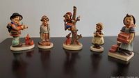 Front view of five Hummel figurines on a table showing different children in various poses and activities.