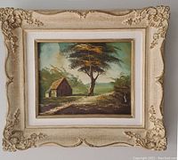 Framed oil painting showing a rustic scene with a house, tree, and pathway in a natural setting.