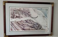 Framed print of a snow leopard in a mountainous snowy setting. Print is matted and framed in dark wood with gold corner accents. Signed and edition number visible.