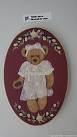 Full view of oval wooden plaque with teddy bear painting on burgundy background featuring floral border.