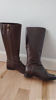 Pair of brown leather knee-high boots standing upright on a wooden floor, showing side profile with zipper and ankle strap details.