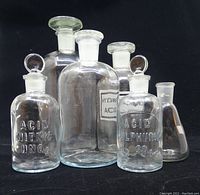 Front view of six antique clear glass laboratory bottles, two with embossed chemical names and one with a paper label