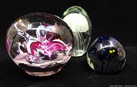 Three art glass paperweights shown together on black background: a floral design paperweight in pink/red, a jellyfish paperweight in oval clear glass, and a round clear glass paperweight with white core.