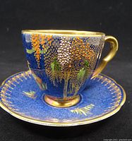 Side angle of hand painted demitasse cup on matching saucer showing Paradise Bird and Tree pattern in cobalt blue base with vibrant multi-colored foliage and birds plus gold gilding.