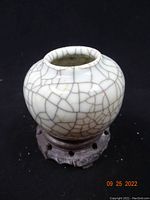 Front angled photo showing small round vase with white crackled glaze on wooden carved stand