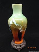 Side view of the Chinese porcelain vase showing the multi-color glaze with green, beige, orange, and black tones and white accents, sitting on a carved wooden base.