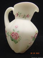 Front angled view of opaque milk glass jug with hand painted pink and green floral designs and curved handle.