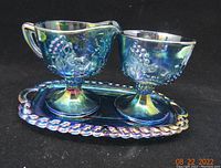Front angled view showing cream pitcher, sugar bowl, and tray with iridescent rich blue carnival glass finish and grape decoration.