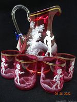 Front-side view of cranberry glass jug with four tumblers, showing white Mary Gregory style painting and gold accents.